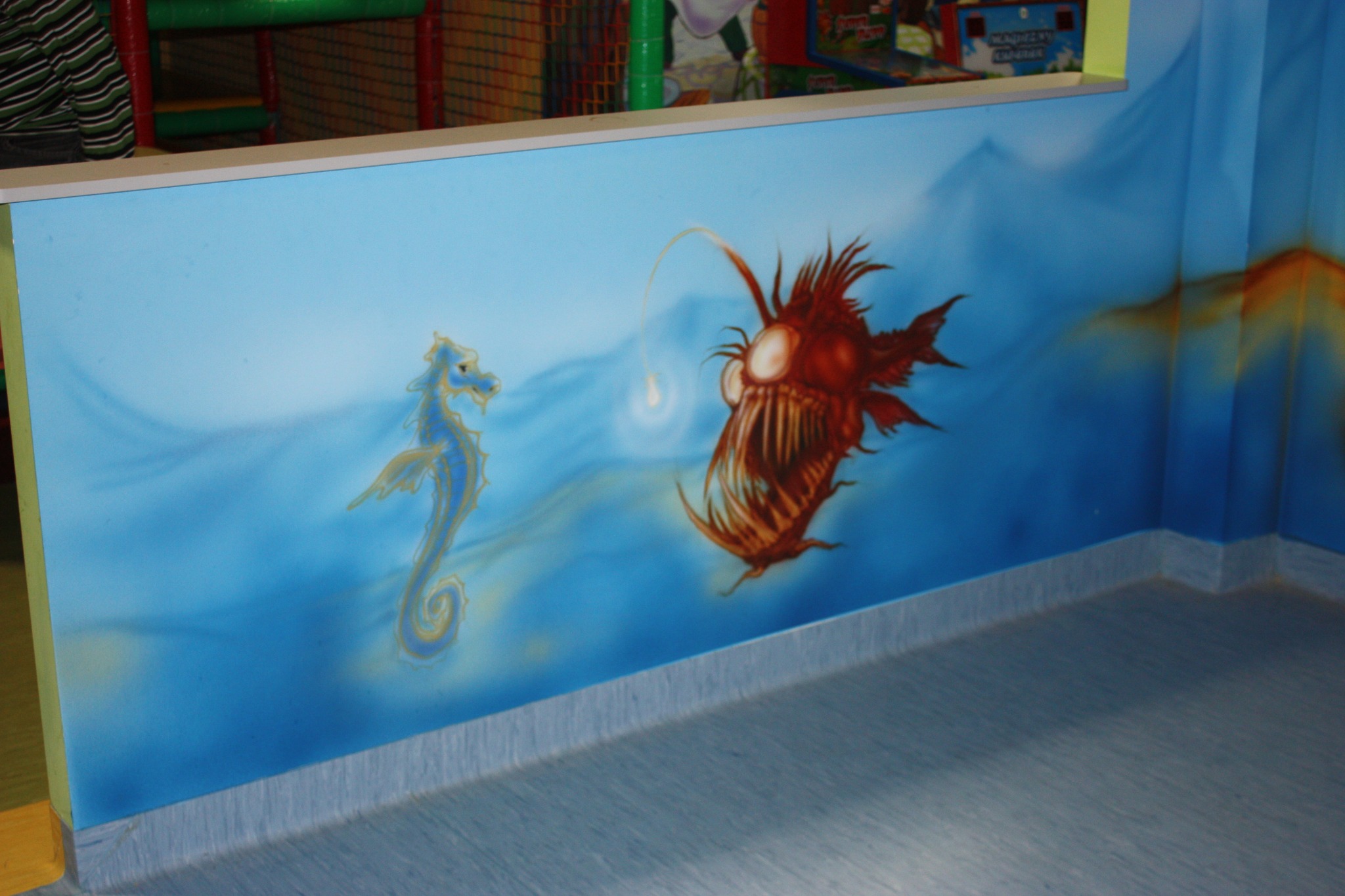 Mural w pokoju dziecięcym przedstawiający podwodny świat z kreskówkowym konikiem morskim i przerażającym żabnicą na błękitnym tle imitującym wodę.