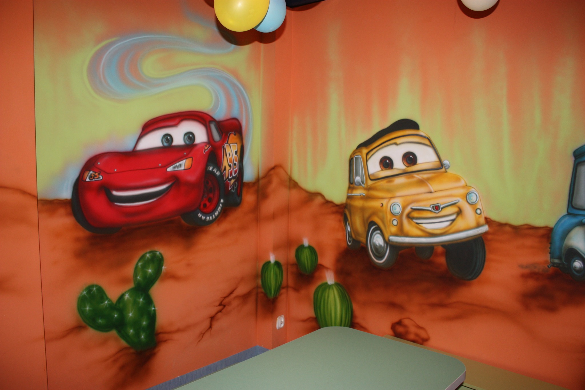 Ściana pokoju dziecięcego z ręcznie malowaną sceną z filmu Auta, z dominującymi postaciami Zygzaka McQueen i Luigi, uzupełniona balonami w pastelowych kolorach.