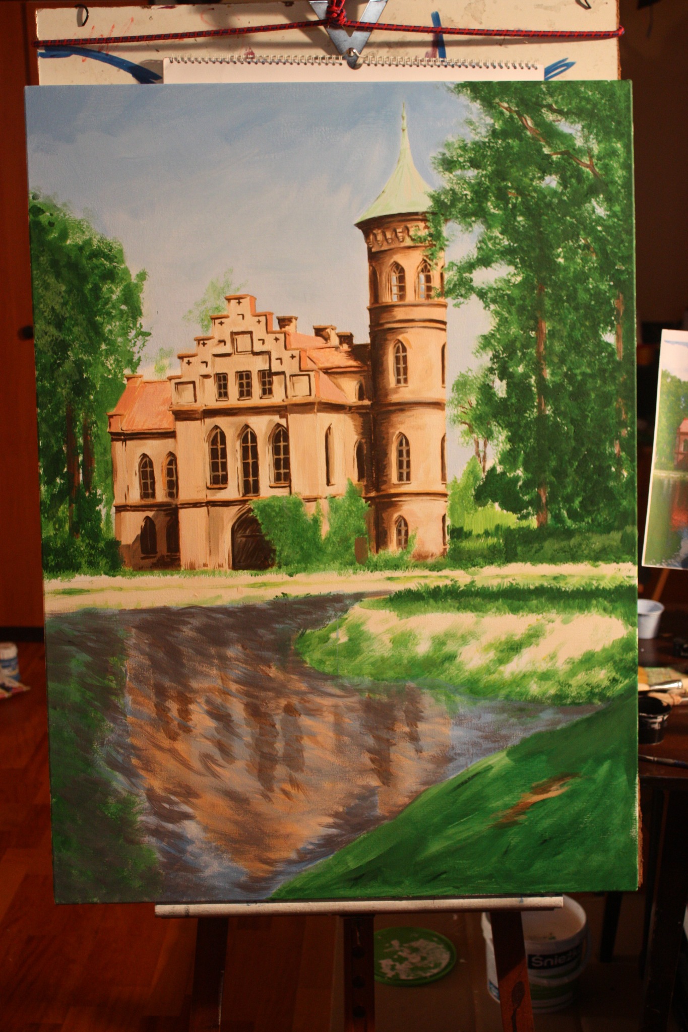 Obraz olejny przedstawiający zamek z wieżą, otoczony zielenią, z odbiciem w wodzie, umieszczony na sztaludze.