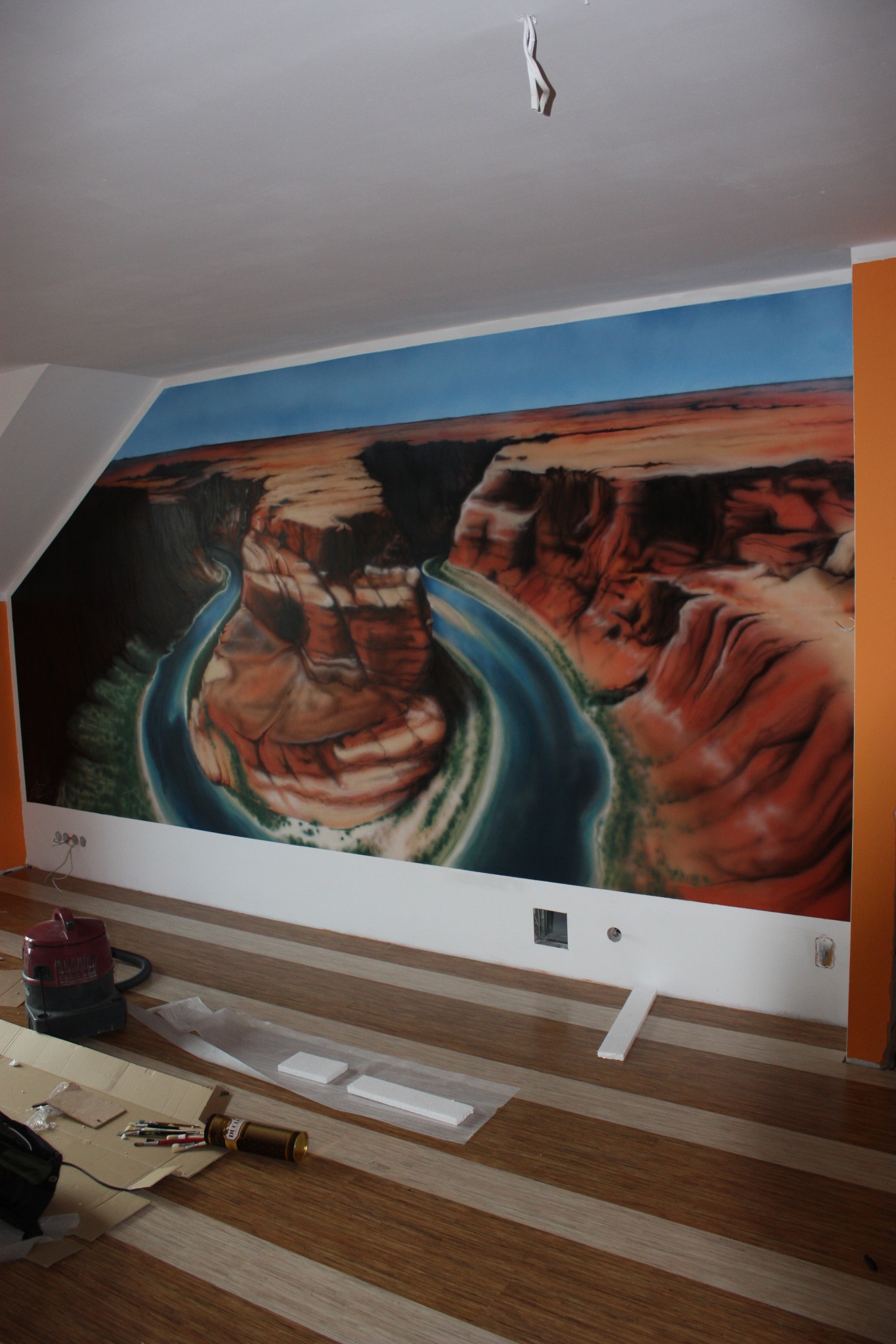 Pejzaż kanionu rzecznego namalowany na ścianie w pokoju, widoczne narzędzia malarskie i odkurzacz, listwa przypodłogowa w trakcie montażu.