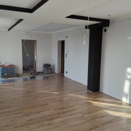 Mir-Bud - Jasne panele podłogowe w nowym wnętrzu z pomalowanymi na szaro ścianami i czarnymi elementami dekoracyjnymi sufitu. Widoczne narzędzia i materiały budowlane.