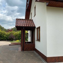 Mir-Bud - Biała elewacja domu z ceglanym podmurówką i zadaszeniem wejścia, widok z boku na tle zieleni i pochmurnego nieba. Detal wykończenia.