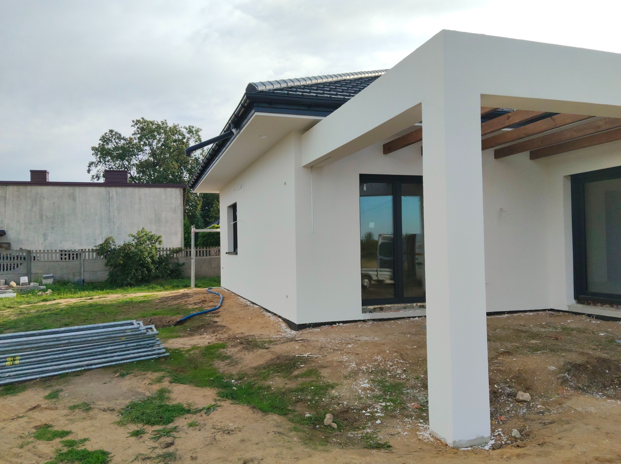 Biały, jednopiętrowy dom z płaskim dachem i przylegającą pergolą. Widoczne rusztowania i teren po budowie. Nowoczesna architektura, jasna elewacja, ciemna dachówka.