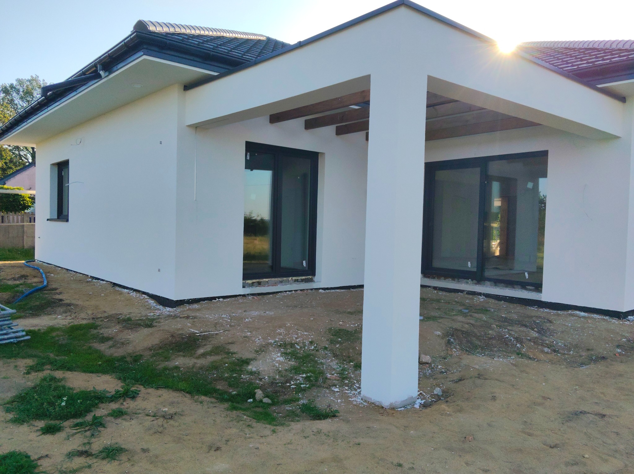 Biały, parterowy dom z płaskim dachem i dużymi oknami, widziany z zewnątrz w słoneczny dzień. Widoczne elementy drewniane pod dachem i świeżo pomalowana elewacja.