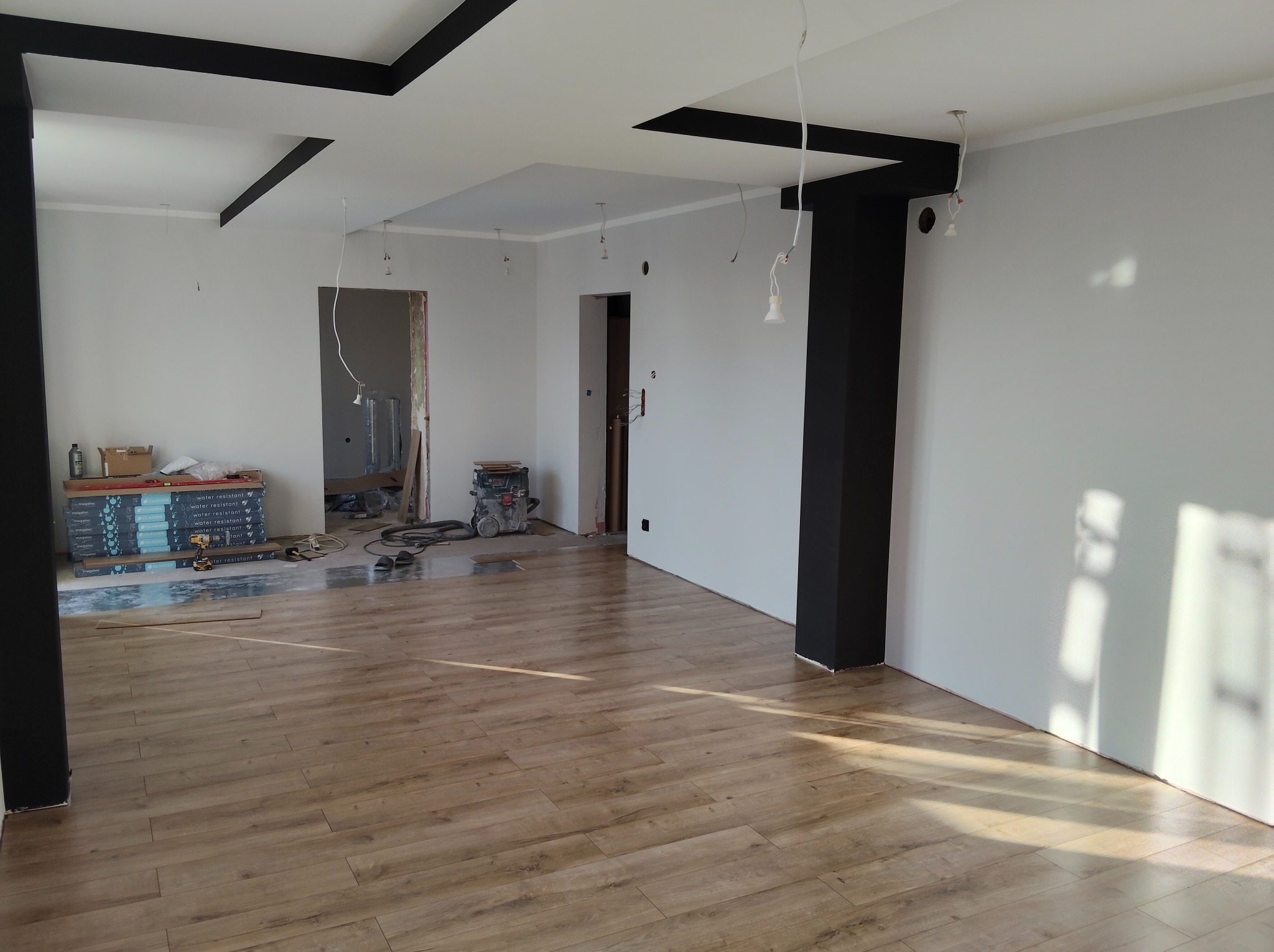 Jasne panele podłogowe w nowym wnętrzu z pomalowanymi na szaro ścianami i czarnymi elementami dekoracyjnymi sufitu. Widoczne narzędzia i materiały budowlane.