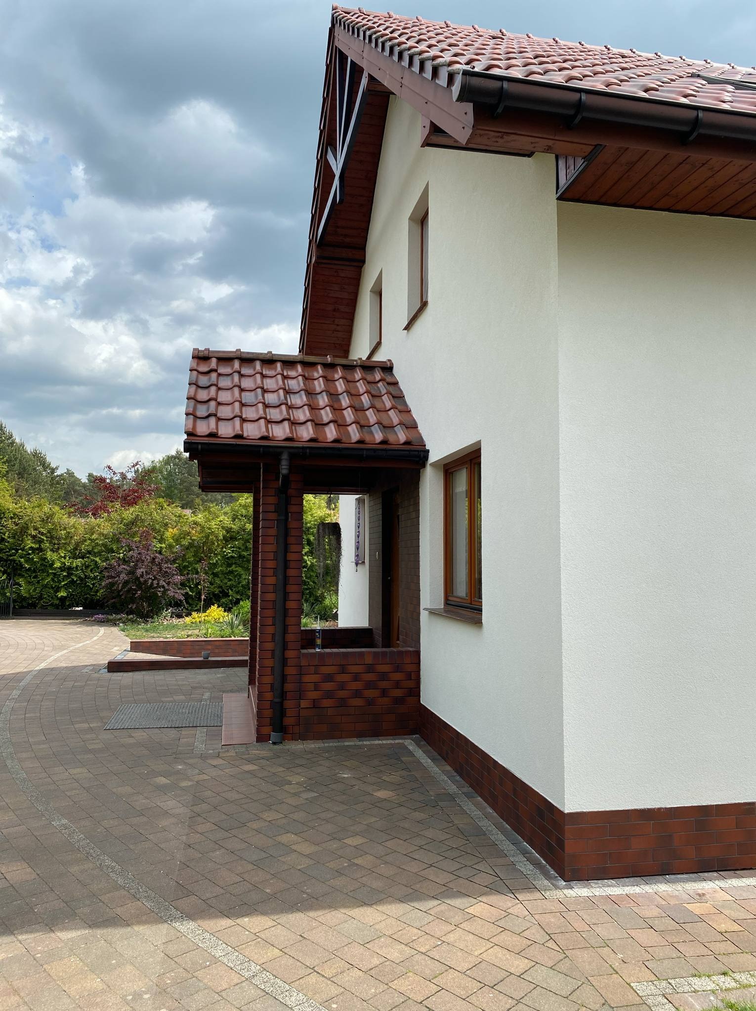Biała elewacja domu z ceglanym podmurówką i zadaszeniem wejścia, widok z boku na tle zieleni i pochmurnego nieba. Detal wykończenia.