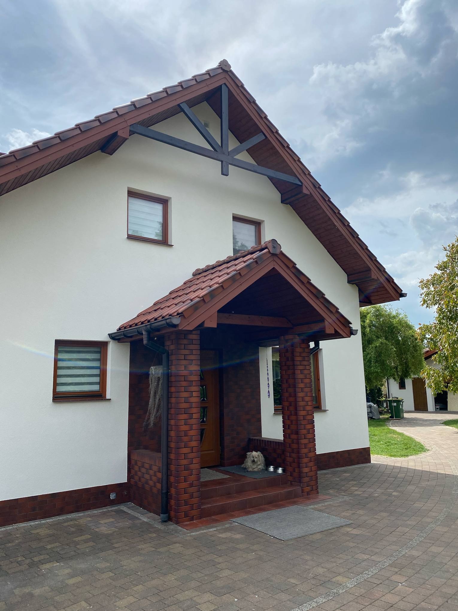 Biały dom jednorodzinny z ceglaną podmurówką i zadaszeniem nad wejściem. Widoczny bruk na podjeździe i fragment ogrodu. Uwagę przykuwa pies leżący na wycieraczce.