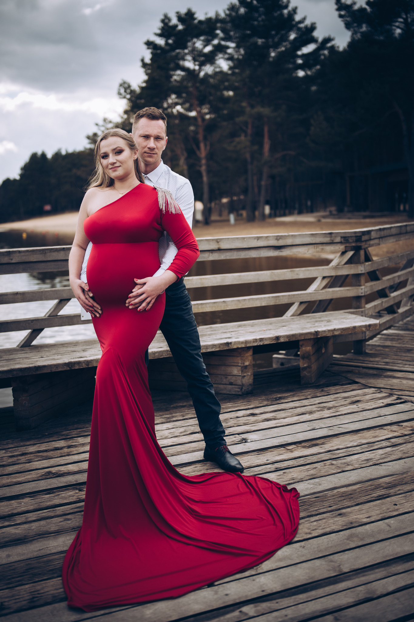 Kobieta w czerwonej sukni ciążowej z partnerem na drewnianym molo, w tle jezioro i las.