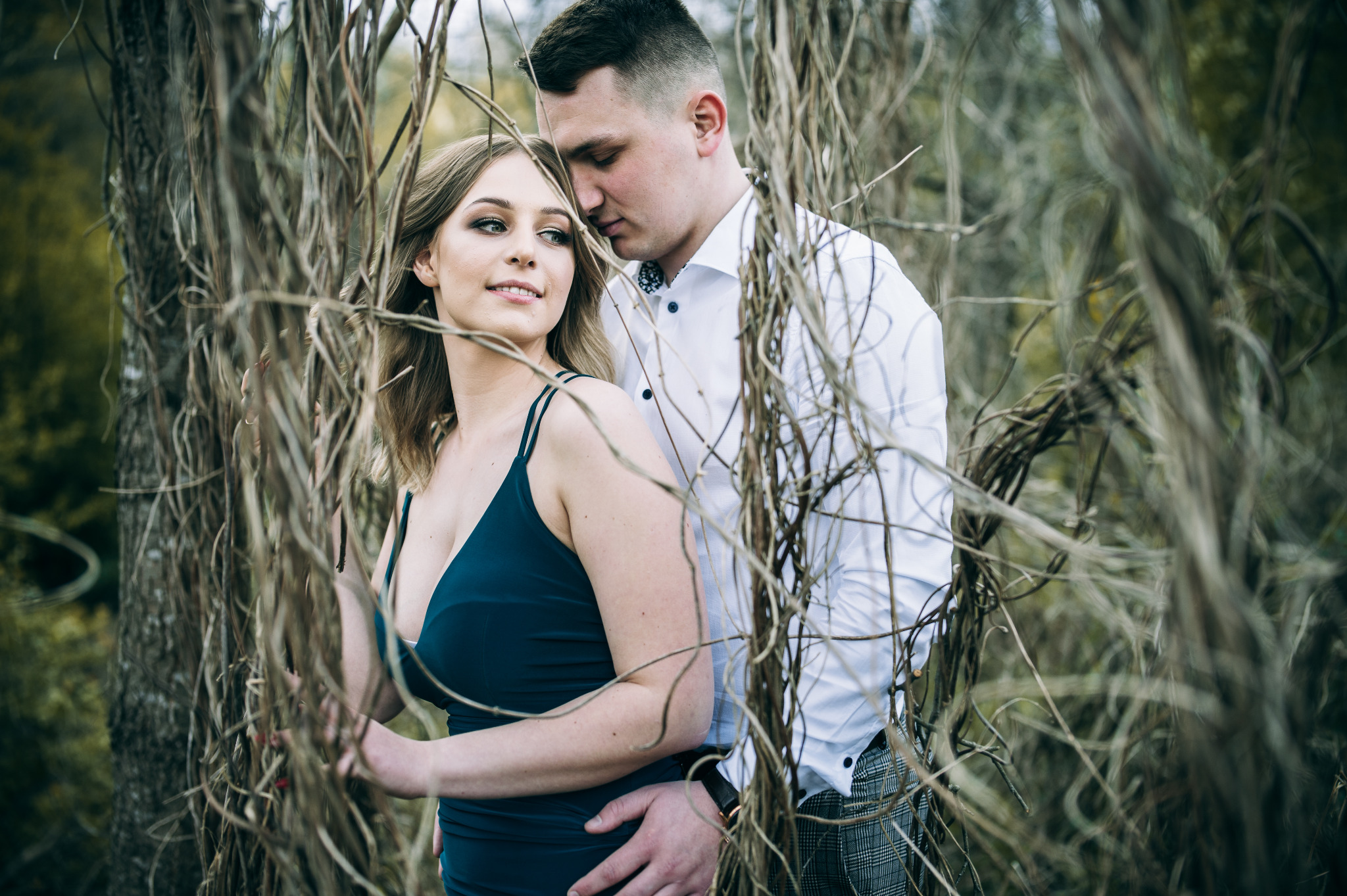 Para obejmująca się pośród suchych gałęzi: kobieta w granatowej sukience, mężczyzna w białej koszuli i spodniach w kratę, subtelne oświetlenie.