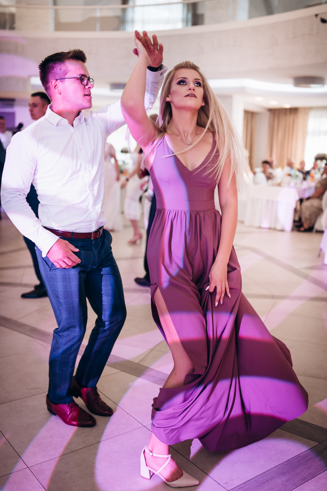 Para tańczy na sali balowej, kobieta w długiej, fioletowej sukni z rozcięciem, mężczyzna w niebieskich spodniach w kratę i białej koszuli, oświetlenie w odcieniach fioletu.