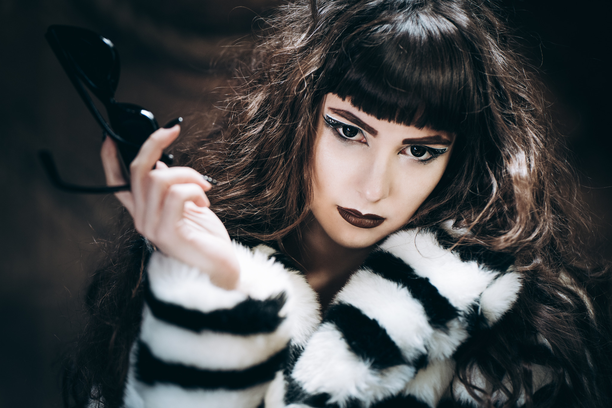 Portret kobiety z mocnym makijażem, w futrze w czarno-białe paski, trzymającej czarne okulary przeciwsłoneczne.