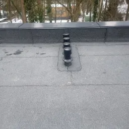 Widok z góry na płaski dach pokryty papą z czterema kominkami wentylacyjnymi, w tle drzewa i zabudowania.