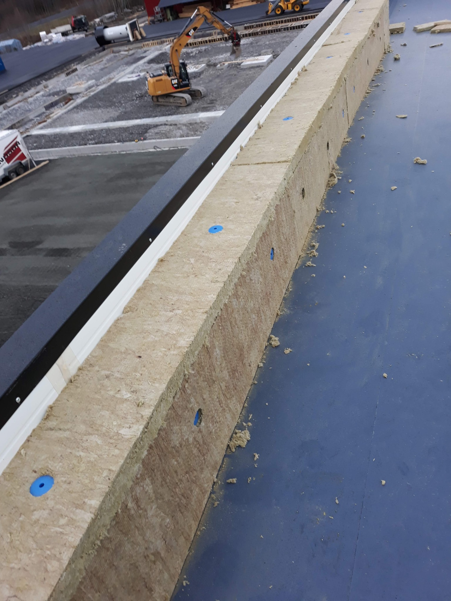 Montaż izolacji termicznej na płaskim dachu z widoczną membraną i elementami mocującymi, w tle teren budowy z koparką.