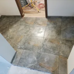 Ułożona podłoga z płytek imitujących marmur w przejściu, widoczny fragment pomieszczenia z żółtym statywem oświetleniowym i zwiniętym kablem w tle.