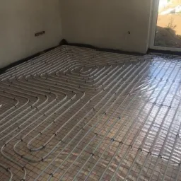 Instalacja ogrzewania podłogowego z rur na siatce, widok z góry na przygotowaną powierzchnię przed wylaniem posadzki, naturalne światło z okna.