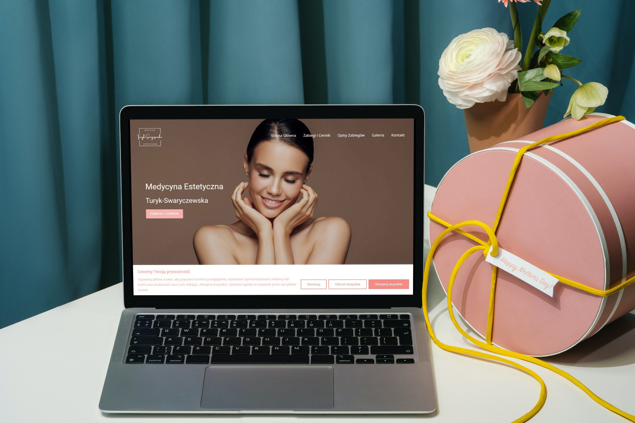 Laptop wyświetlający stronę internetową gabinetu medycyny estetycznej z uśmiechniętą kobietą, obok różowe pudełko prezentowe z żółtą wstążką i kartką z napisem 'Happy Mother's Day'.