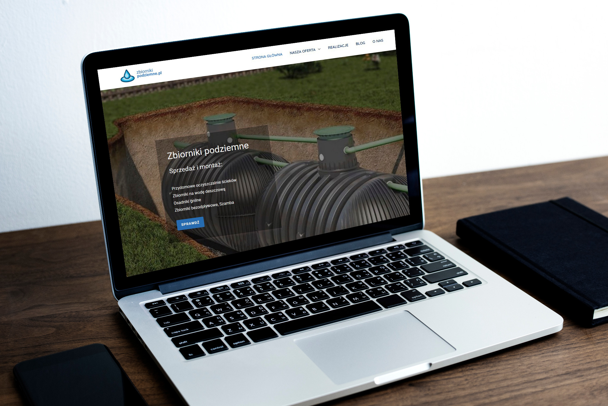 Laptop wyświetla stronę internetową z wizualizacją podziemnych zbiorników na wodę deszczową. Obok leży czarny notatnik i smartfon na drewnianym biurku.