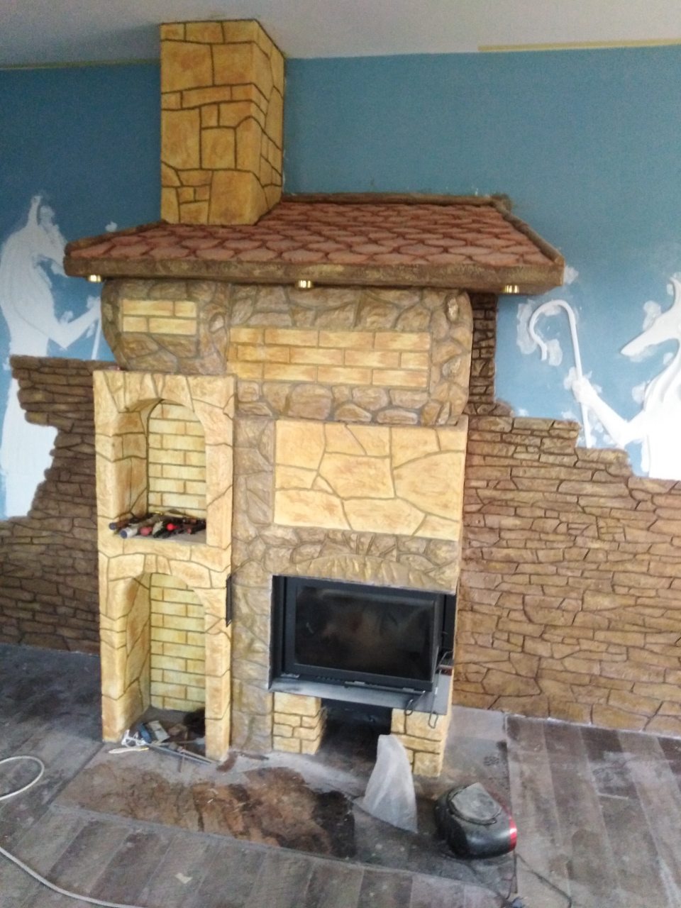 Ozdobny kominek z imitacją kamienia i cegły, z wkładem kominkowym, na tle niebieskiej ściany z białymi konturami postaci, widoczne niedokończone prace wykończeniowe.