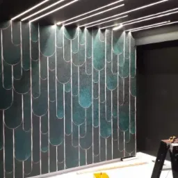 Ściana z dekoracyjną, zielono-złotą tapetą o geometrycznym wzorze, oświetlona paskami LED w suficie, z drabinką malarską w prawym dolnym rogu.