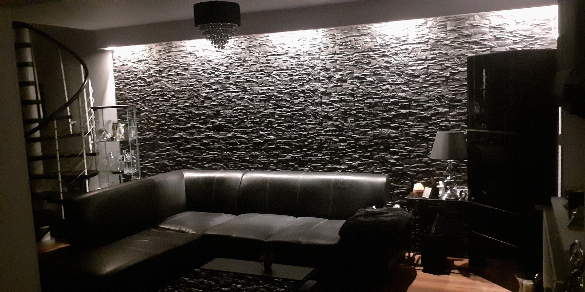 Salon z ciemną, kamienną ścianą podświetloną od góry, czarną skórzaną sofą i spiralnymi schodami w tle.