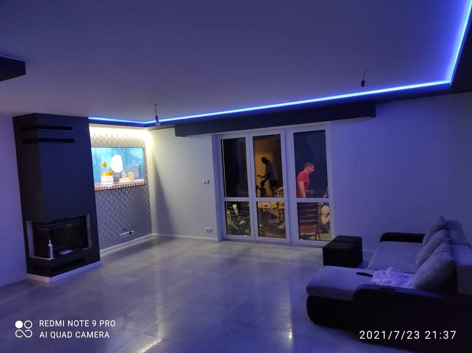 Nowoczesny salon z podświetlanym sufitem LED, kominkiem obudowanym szarą płytą i akwarium w ścianie. W tle widoczne okno balkonowe i osoby wewnątrz.