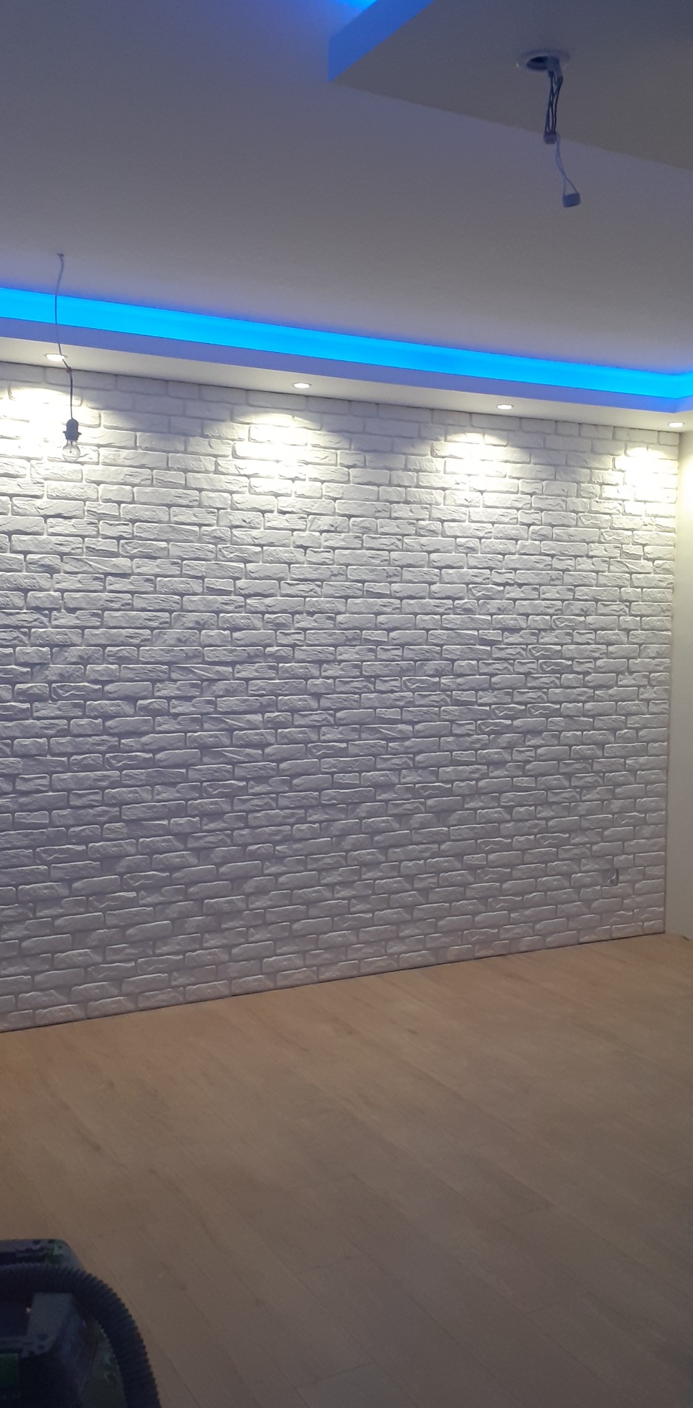 Ściana wykończona białymi panelami imitującymi cegłę, oświetlona punktowo i listwą LED w suficie, z widoczną instalacją elektryczną w suficie.