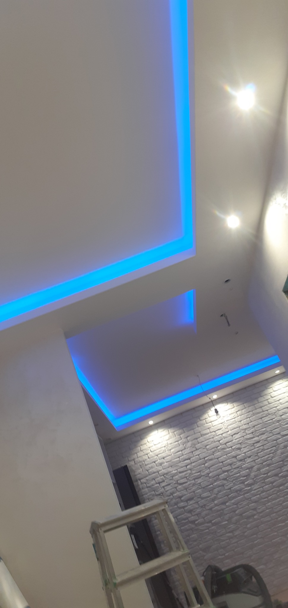 Nowoczesny sufit podwieszany z oświetleniem LED w kolorze niebieskim, ściana z dekoracyjnej cegły, drabina w rogu pokoju.