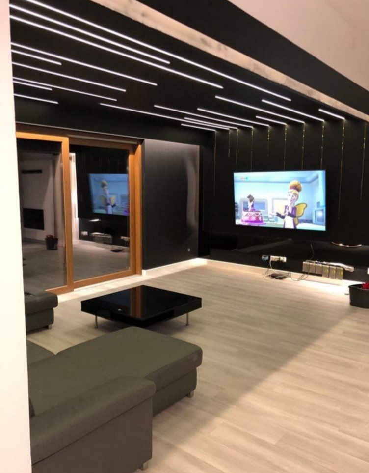 Nowoczesny salon z czarną ścianą, podświetlanym sufitem, telewizorem i panelami podłogowymi w odcieniach szarości, widoczne meble wypoczynkowe.