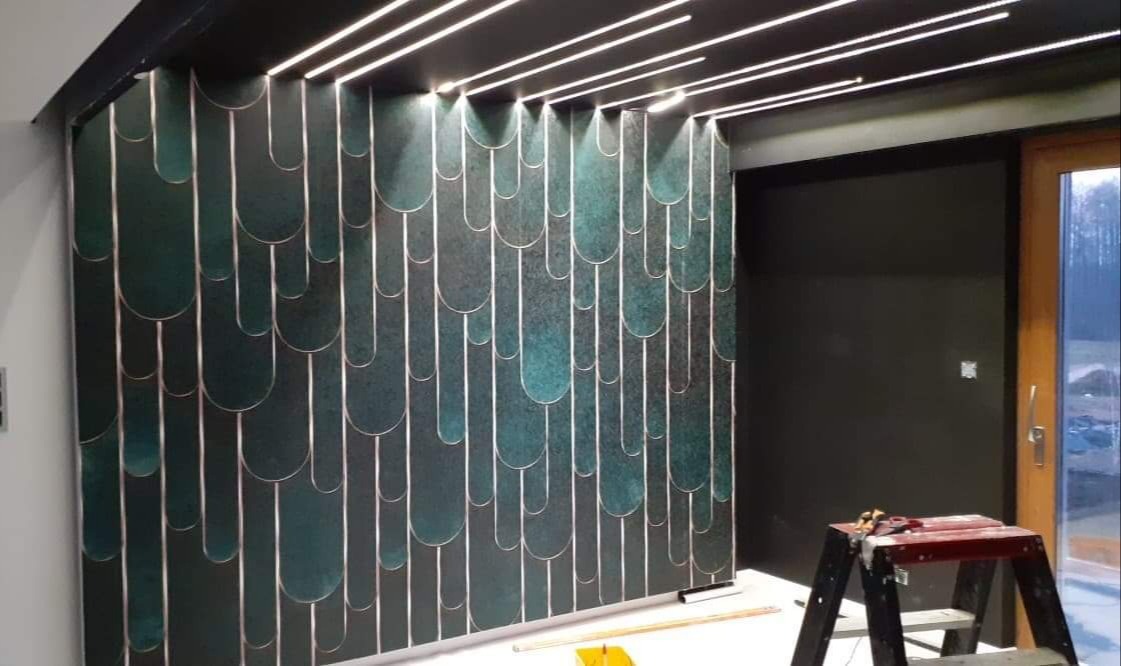 Ściana z dekoracyjną, zielono-złotą tapetą o geometrycznym wzorze, oświetlona paskami LED w suficie, z drabinką malarską w prawym dolnym rogu.