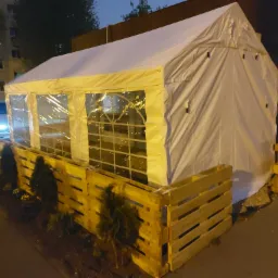 Biały namiot z przezroczystymi ścianami, otoczony drewnianym płotem z palet i małymi drzewkami, oświetlony nocą, z widocznym zaparkowanym samochodem w tle.