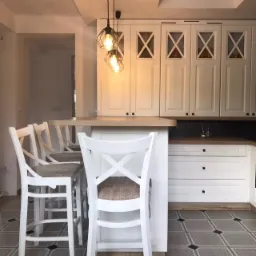 Jasna kuchnia z białymi frontami, wyspą i hokerami w stylu rustykalnym, oświetlona trzema wiszącymi lampami z czarnymi geometrycznymi kloszami.