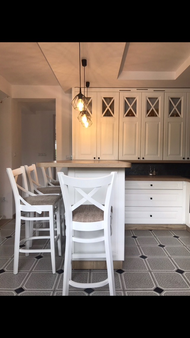 Jasna kuchnia z białymi frontami, wyspą i hokerami w stylu rustykalnym, oświetlona trzema wiszącymi lampami z czarnymi geometrycznymi kloszami.