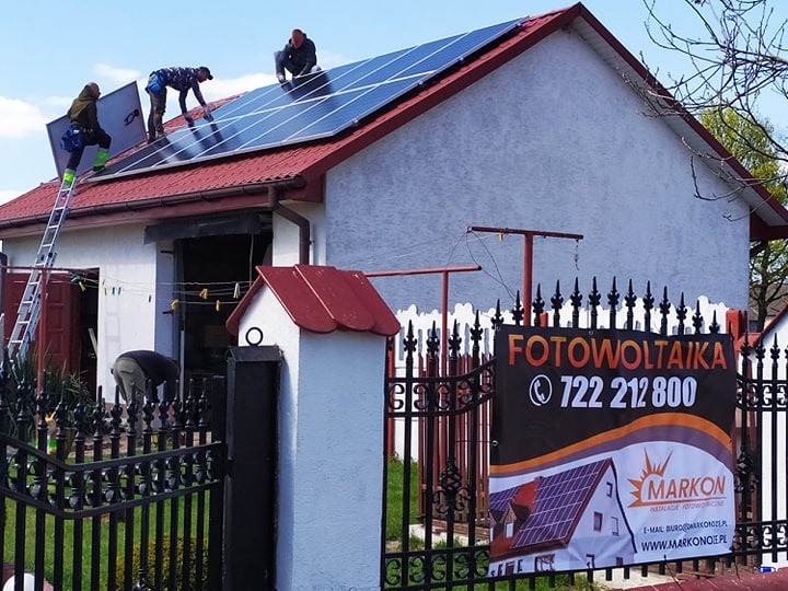 Montaż paneli fotowoltaicznych na dachu domu jednorodzinnego przez ekipę monterów, widoczna drabina, banner reklamowy firmy instalacyjnej przy ogrodzeniu.