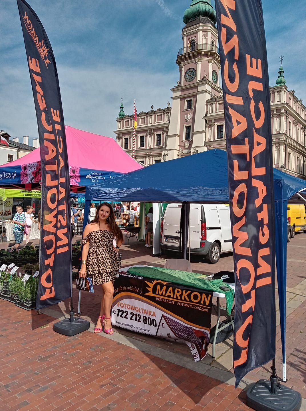 Stoisko firmy Markon na targach, oferującej instalacje fotowoltaiczne. Widoczna kobieta w sukience w panterkę, flagi reklamowe z napisem 'Fotowoltaika', baner z numerem telefonu i logo firmy na tle...