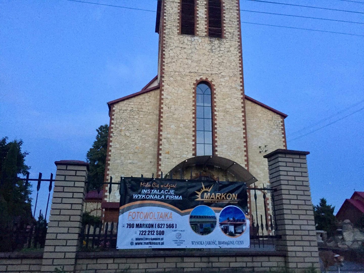 Baner reklamowy firmy instalującej systemy fotowoltaiczne, umieszczony na ogrodzeniu przed budynkiem kościoła z kamienia.
