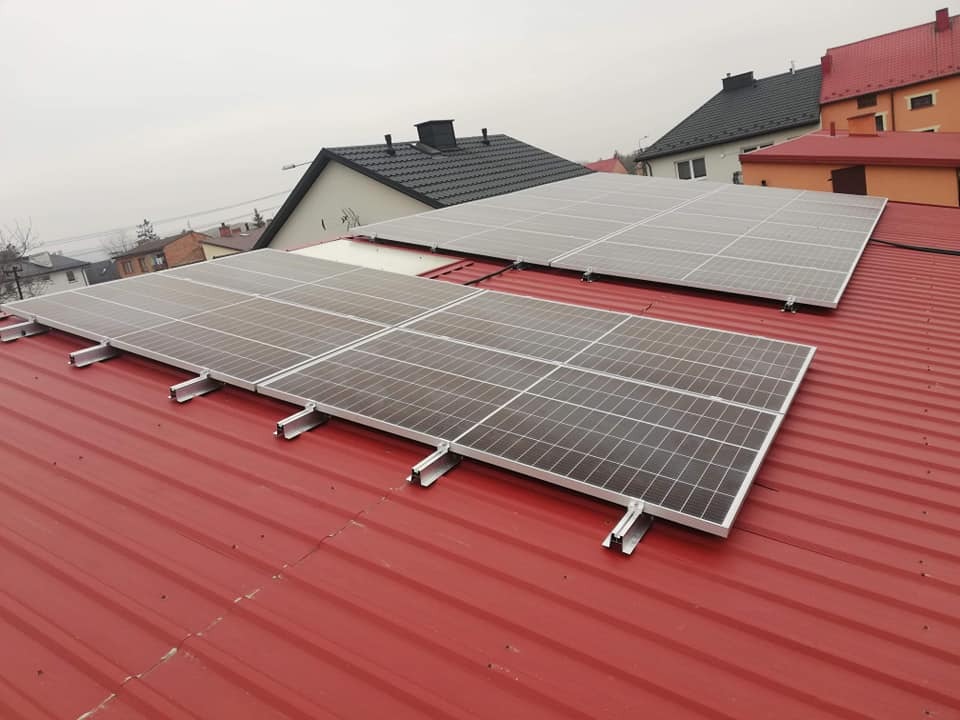 Panele słoneczne zamontowane na czerwonym blaszanym dachu domu jednorodzinnego, widoczne metalowe uchwyty mocujące.