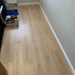 Jasne panele podłogowe ułożone wzdłuż ściany z białą listwą przypodłogową, widoczna skrzynka z narzędziami i niebieska bluza. Grzejnik na ścianie w tle.