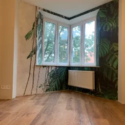 Pokój po remoncie z drewnianą podłogą, ścianami z motywem roślinnym i białym grzejnikiem pod oknem.