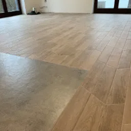 Podłoga wyłożona dwoma rodzajami płytek: imitacja drewna ułożona pod kątem oraz szare, matowe płytki. Widoczne okna balkonowe i gniazdka elektryczne na ścianie.