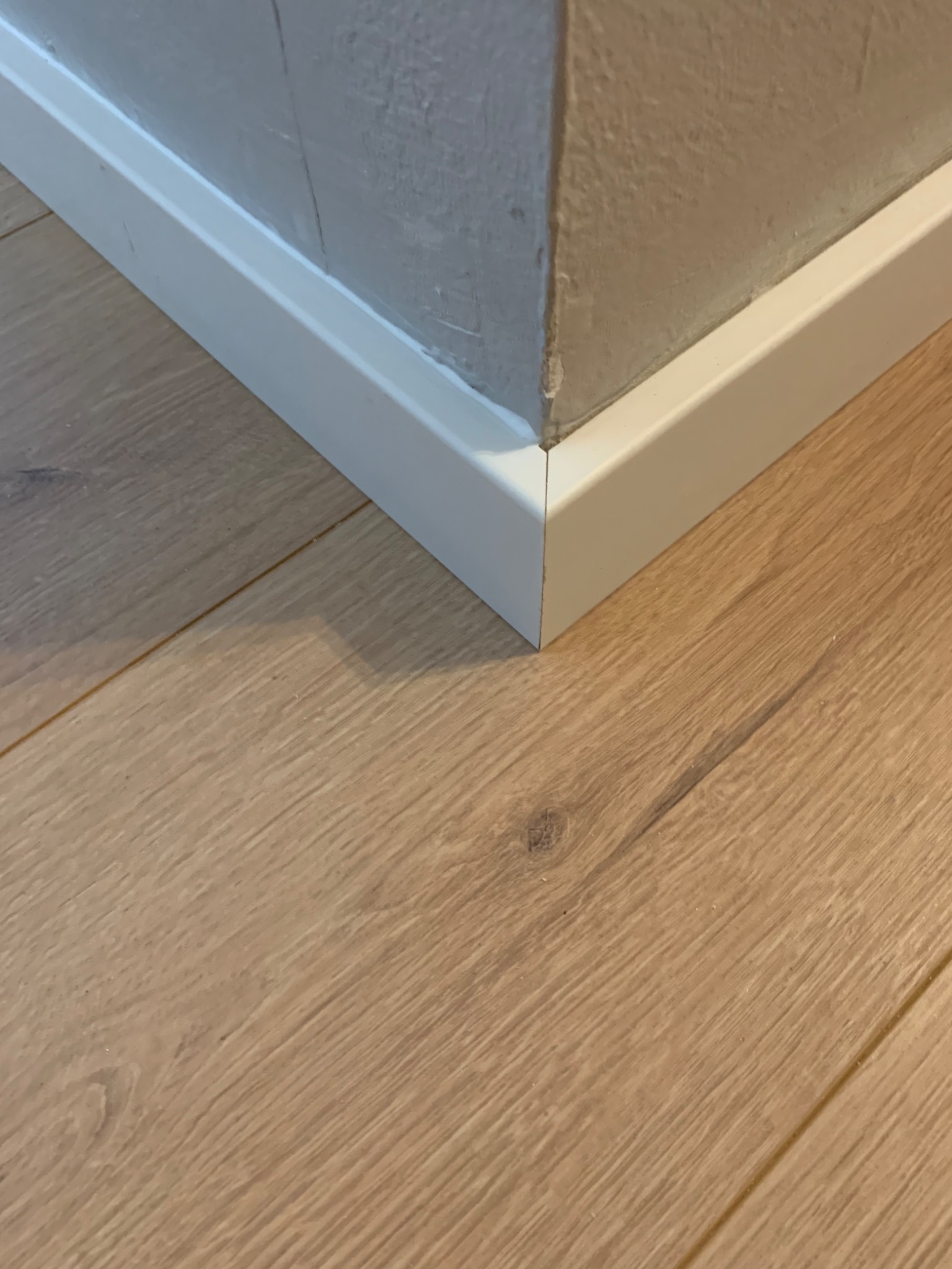 Fragment narożnika pokoju po montażu przypodłogowych listew MDF w kolorze białym, widoczne jasne panele podłogowe i szara ściana.