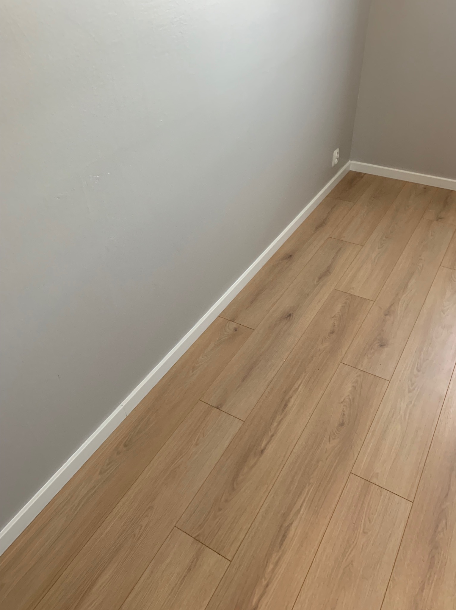 Ułożone panele podłogowe w rogu pomieszczenia, widoczne białe listwy przypodłogowe i szara ściana z gniazdkiem elektrycznym