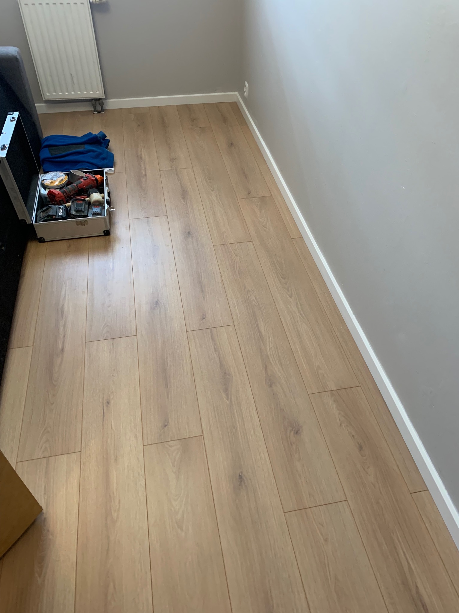 Jasne panele podłogowe ułożone wzdłuż ściany z białą listwą przypodłogową, widoczna skrzynka z narzędziami i niebieska bluza. Grzejnik na ścianie w tle.