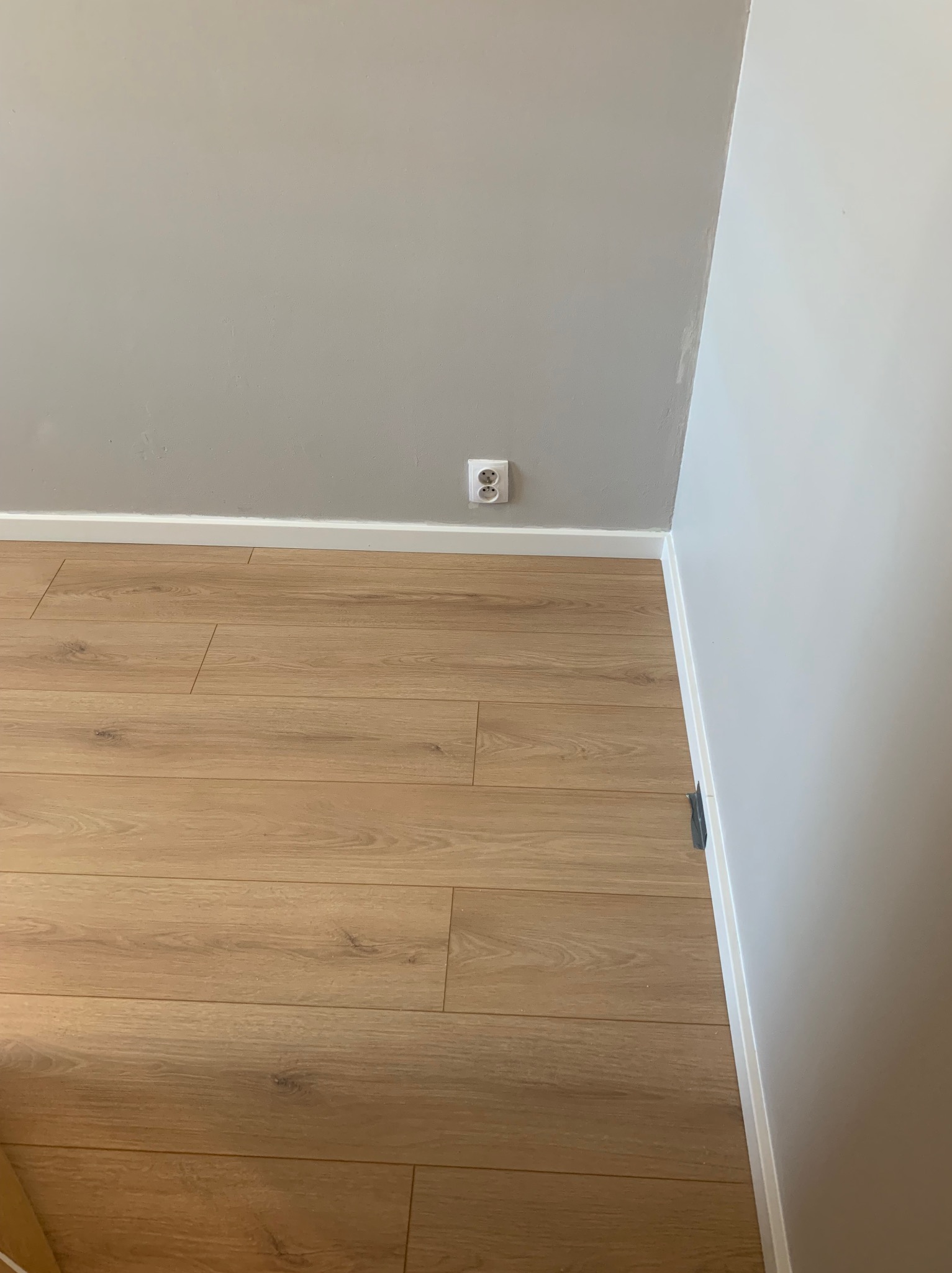 Jasne panele podłogowe ułożone wzdłuż szarej ściany z białym gniazdkiem elektrycznym i białą listwą przypodłogową.