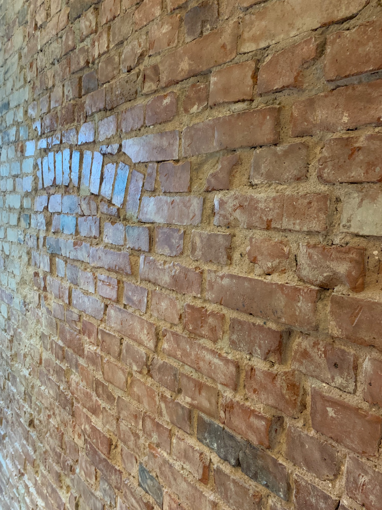 Murek z czerwonej cegły z wyraźnymi spoinami i fragmentami pobielenia, sfotografowany pod kątem, z widocznymi refleksami światła na niektórych cegłach.