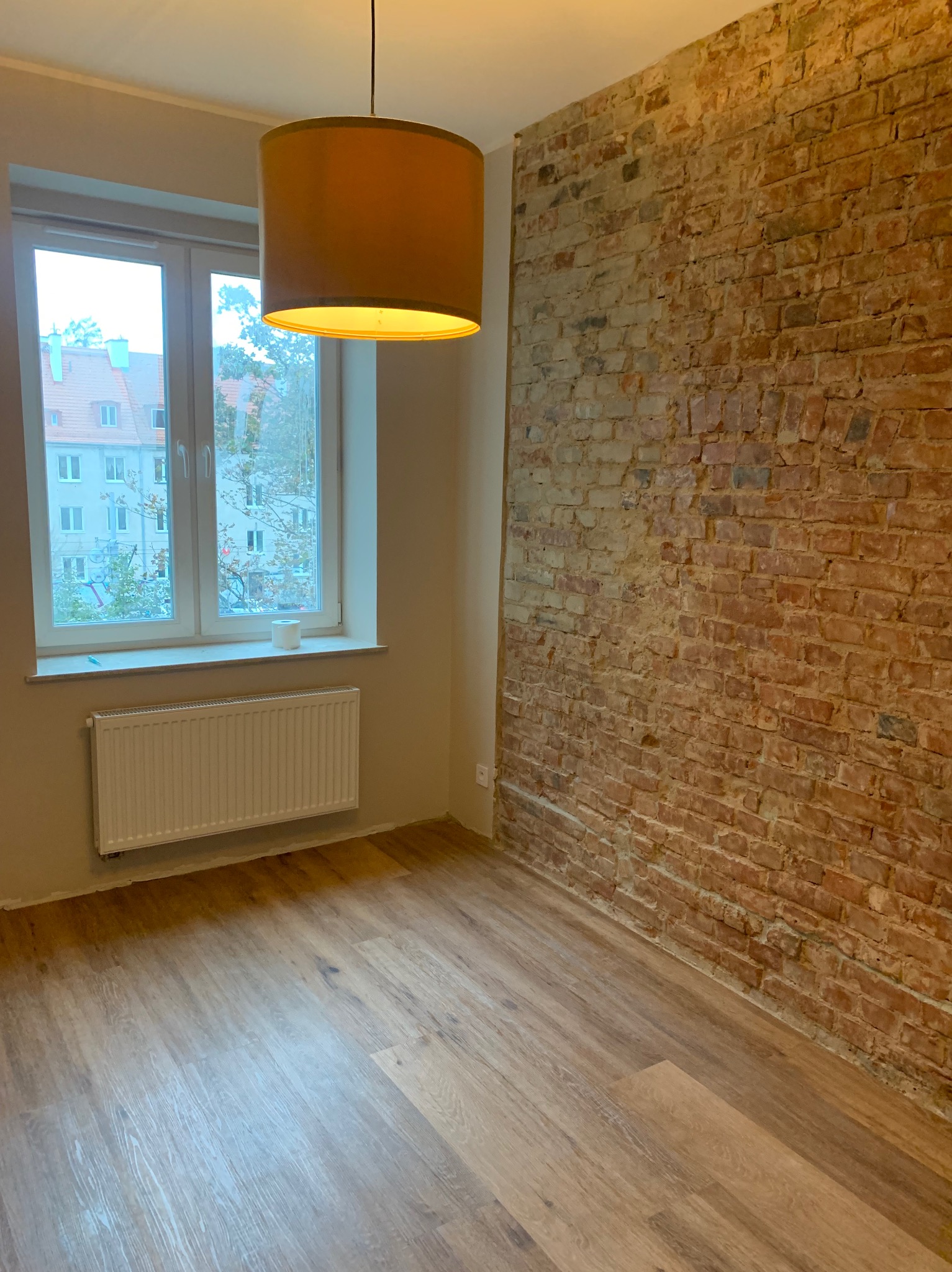 Pokój po remoncie z panelami podłogowymi, odsłoniętą ścianą z czerwonej cegły i wiszącą lampą z abażurem w kolorze musztardowym.