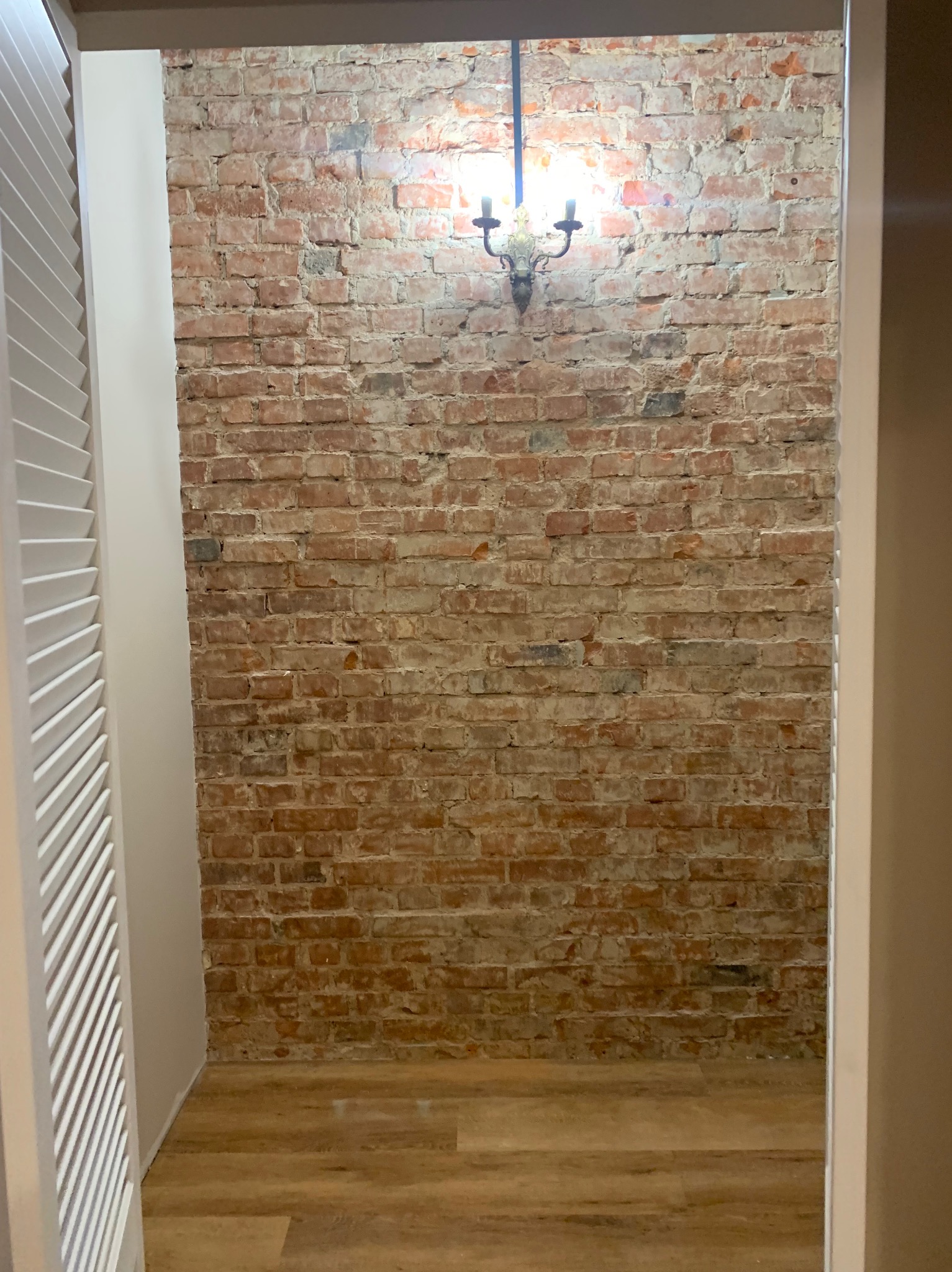 Wnętrze z odsłoniętą czerwoną cegłą na ścianie, oświetloną przez żyrandol w stylu retro, widoczne jasne panele podłogowe i fragment białych żaluzji po lewej stronie.