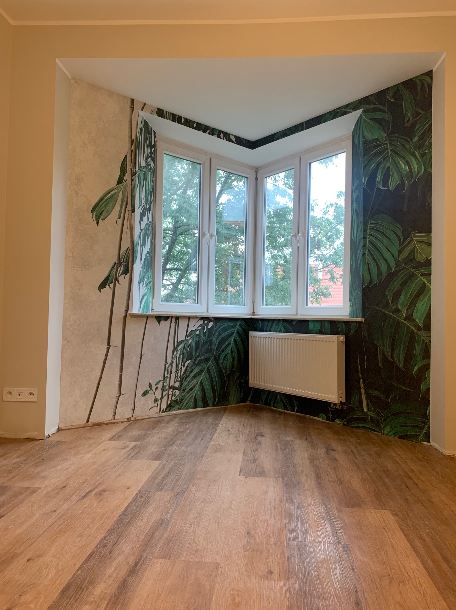 Pokój po remoncie z drewnianą podłogą, ścianami z motywem roślinnym i białym grzejnikiem pod oknem.