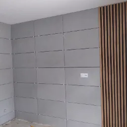 Ściana wyłożona szarymi, poziomymi panelami imitującymi beton, oddzielonymi fugami z krzyżykami dystansowymi, obok pionowy panel dekoracyjny z drewnianych listew, widoczne gniazdko elektryczne.