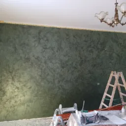 Ściana w pokoju po nałożeniu dekoracyjnej, zielonej farby ze wzorem, zabezpieczona taśmą malarską przy suficie, widoczne drabiny i narzędzia malarskie na folii ochronnej.