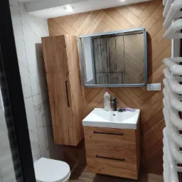 Nowoczesna łazienka z drewnianą ścianą ułożoną w jodełkę, wiszącą szafką z umywalką, lustrem w metalowej ramie i białym grzejnikiem drabinkowym.